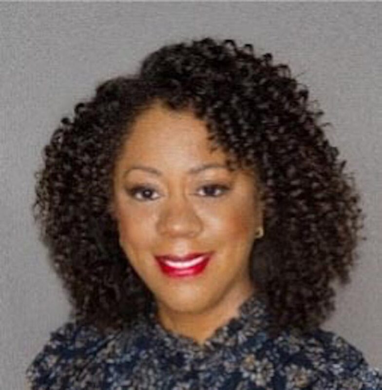 Person with curly hair, smiling, wearing a patterned top; gray background.