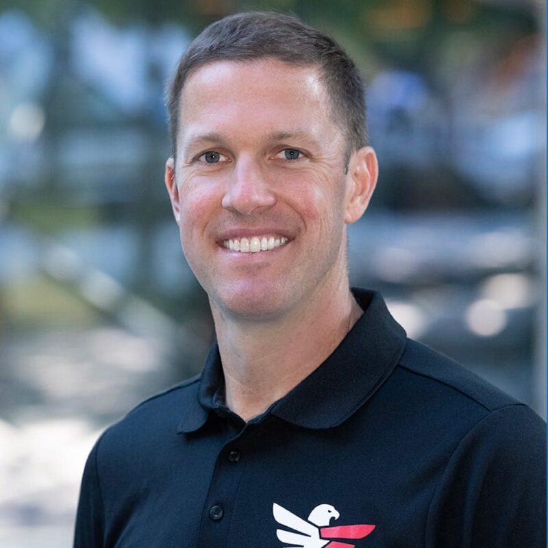 A smiling man wearing a black polo shirt with a logo stands outdoors with a blurred background.