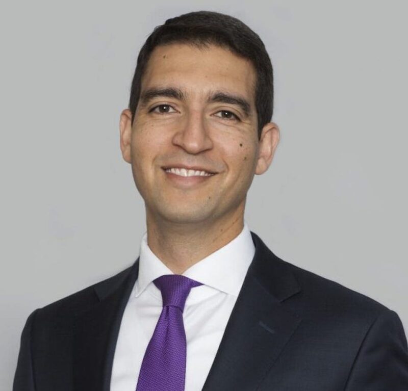 A smiling man in a dark suit, white shirt, and purple tie against a plain gray background.