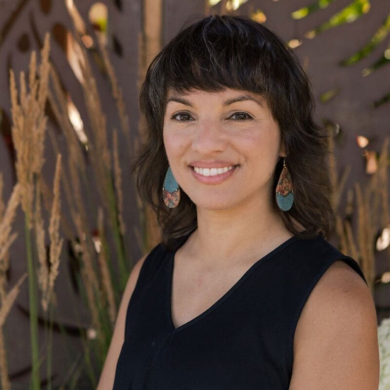 Smiling person with shoulder-length hair, wearing a sleeveless black top and blue earrings, standing outside with tall grass in the background.