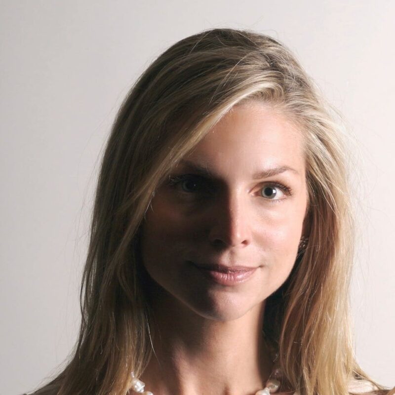 A person with long blonde hair and a light complexion, wearing a pearl necklace, smiling subtly against a white background.