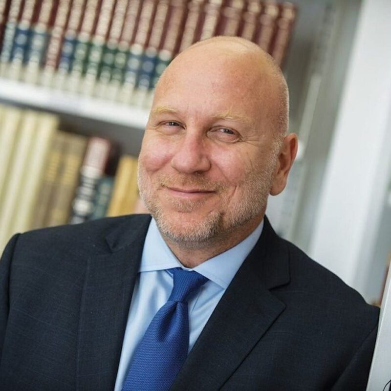 Smiling bald man in a suit and blue tie stands in front of bookshelves.