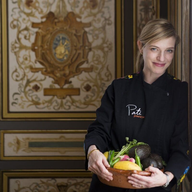 Chef in a dark uniform holds a bowl of fresh vegetables, smiling. Ornate gold and brown patterned wall in the background.