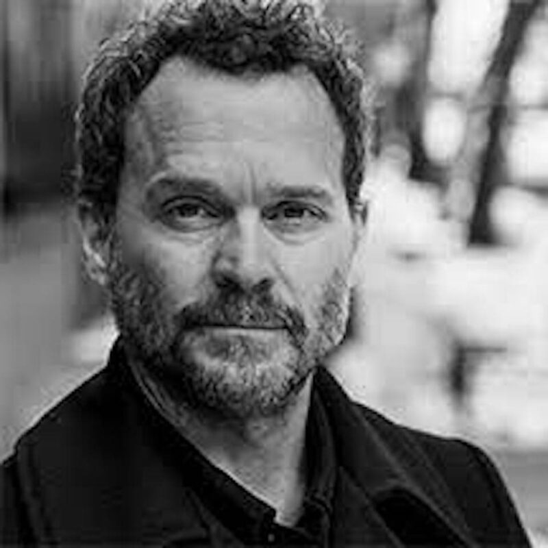 Black and white photo of a man with a beard, wearing a dark coat, looking directly at the camera, with blurred trees in the background.