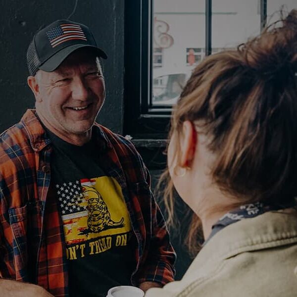Man in a plaid shirt and baseball cap talks smilingly with a woman in a denim jacket in a dimly lit room.