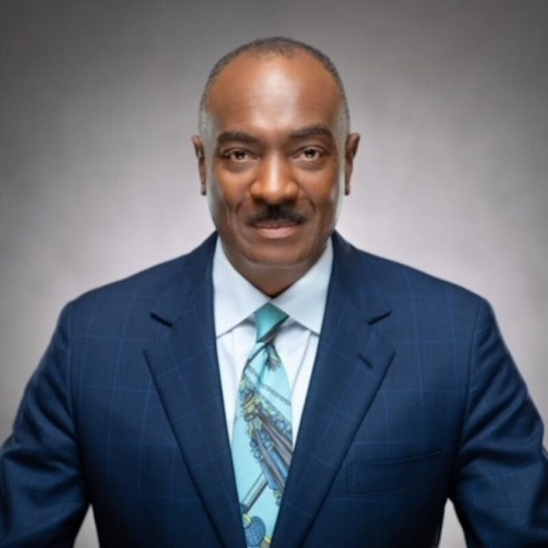 A man in a blue suit and patterned tie, with a neutral expression, stands against a gray background.
