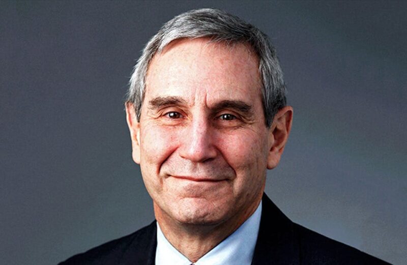 A person with gray hair, wearing a suit and tie, smiles against a gray background.