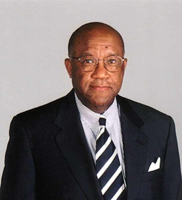 A man in a suit with a striped tie stands against a plain background, wearing glasses.