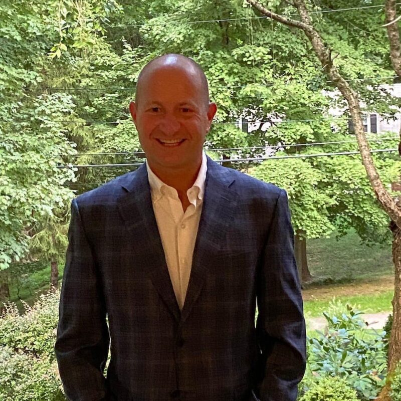 Man in a dark suit stands smiling in front of a lush, green outdoor background with trees and bushes.