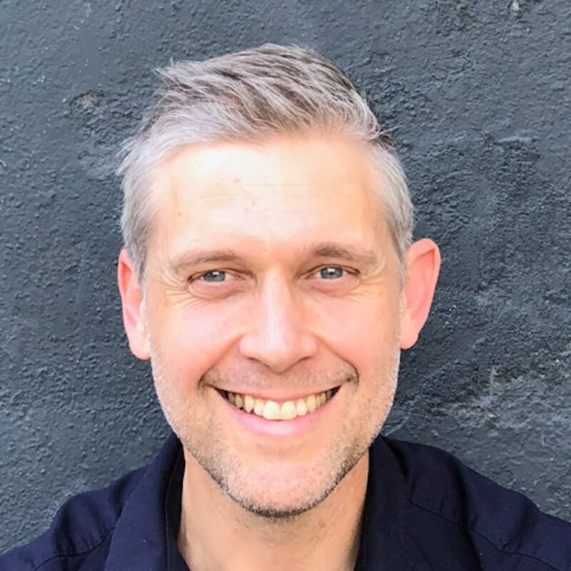 Smiling man with short gray hair and a black shirt against a dark textured wall.