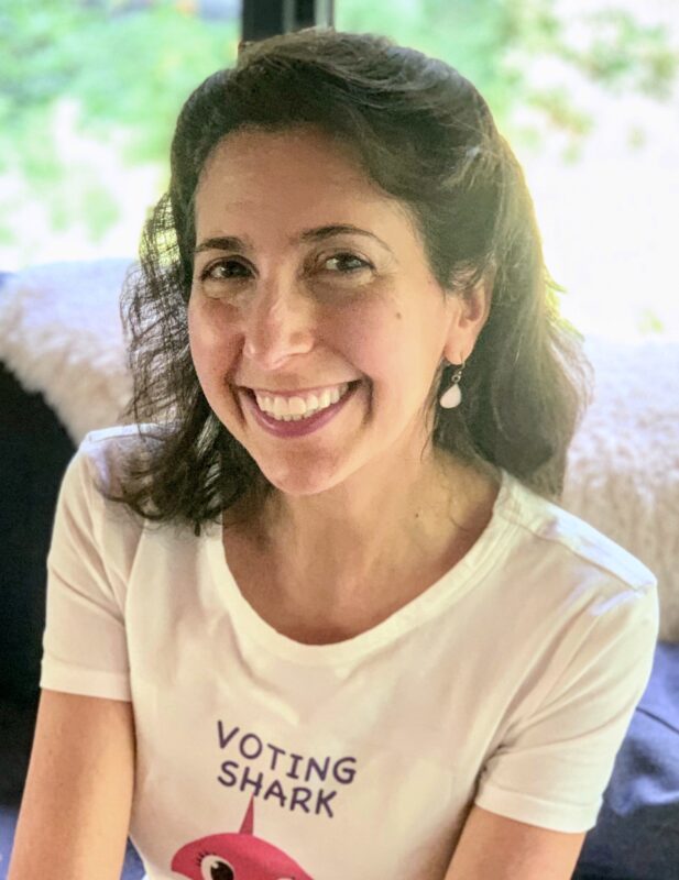 Woman smiling, wearing a t-shirt with "Voting Shark" text and an illustration of a shark. She sits indoors with a blurred background.