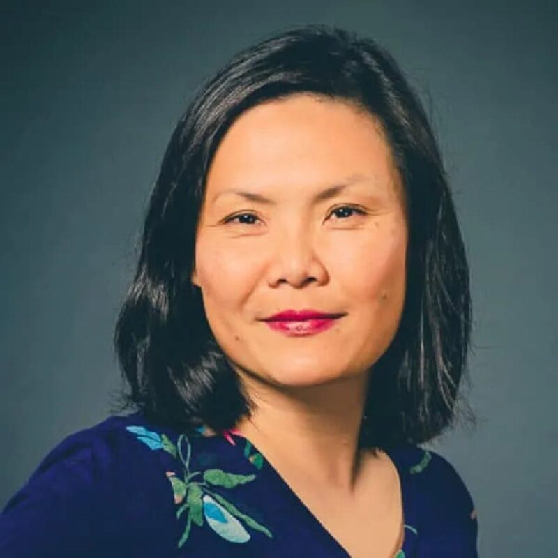 Person with shoulder-length dark hair, wearing a floral-patterned shirt, against a dark background.