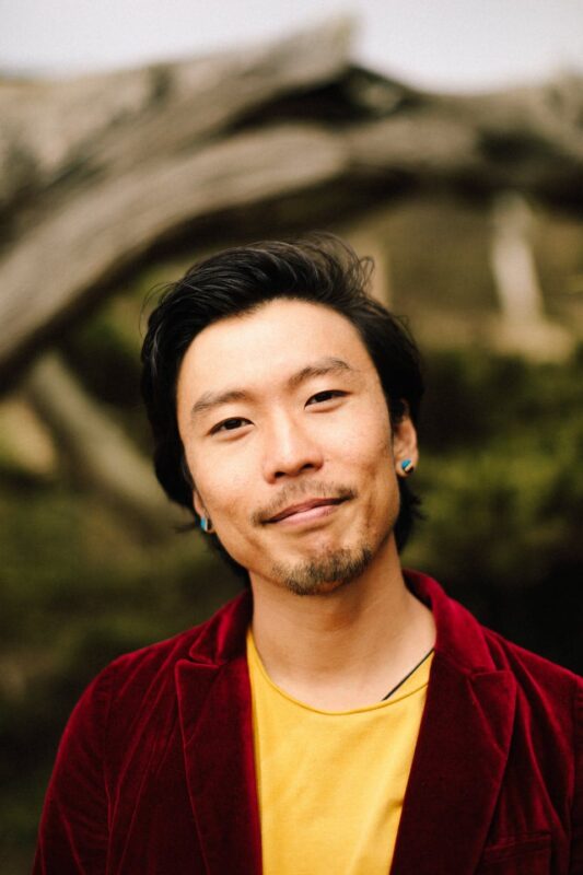 A person wearing a red jacket and yellow shirt smiles slightly, standing outdoors with blurred greenery and branches in the background.