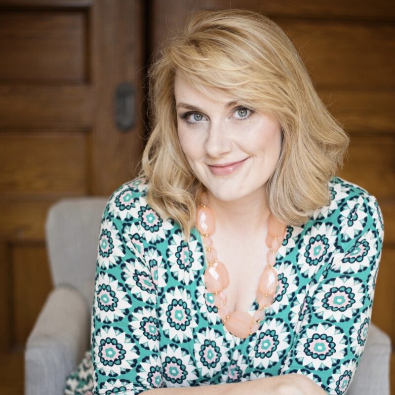 Smiling woman with shoulder-length blonde hair, wearing a patterned dress and a chunky peach necklace, sitting indoors with a wooden background.