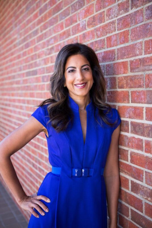 Person in a blue dress leaning against a red brick wall, smiling with hand on hip.