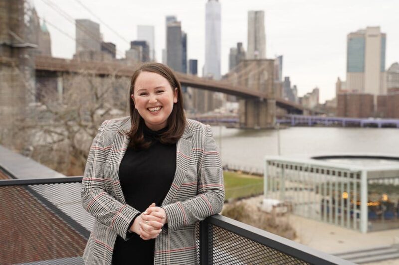 Woman smiling, standing on a balcony with the Brooklyn Bridge and Manhattan skyline in the background, on a cloudy day.