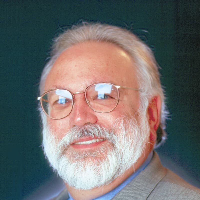 Older man with glasses, white beard, and gray hair, wearing a suit, against a dark background.