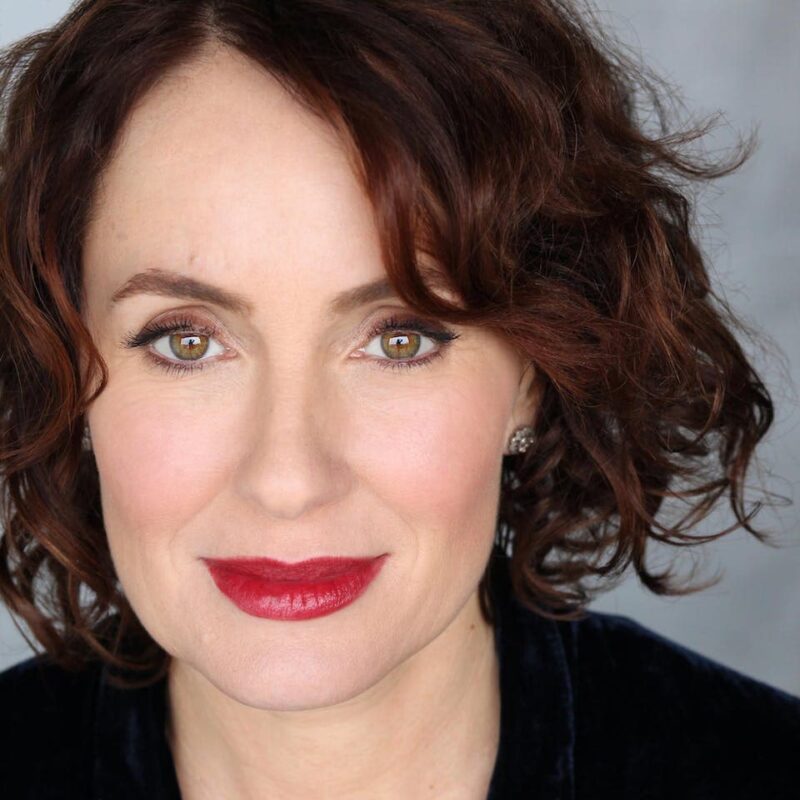 A person with short, curly brown hair and red lipstick smiles softly, wearing a dark top, against a neutral background.