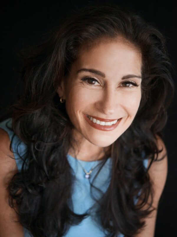 Woman with long, curly dark hair smiling, wearing a light blue top and necklace, against a dark background.