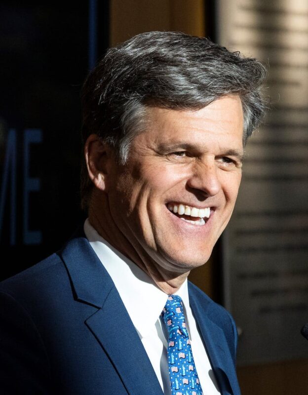 Smiling man in a suit with a blue patterned tie, standing indoors.
