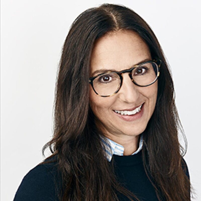 Smiling person with long dark hair, wearing glasses and a dark top over a striped shirt, against a plain white background.