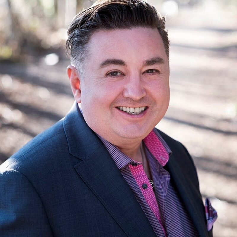 Man smiling outdoors in a suit jacket with a pink shirt, standing on a path with blurred sunlight and trees in the background.