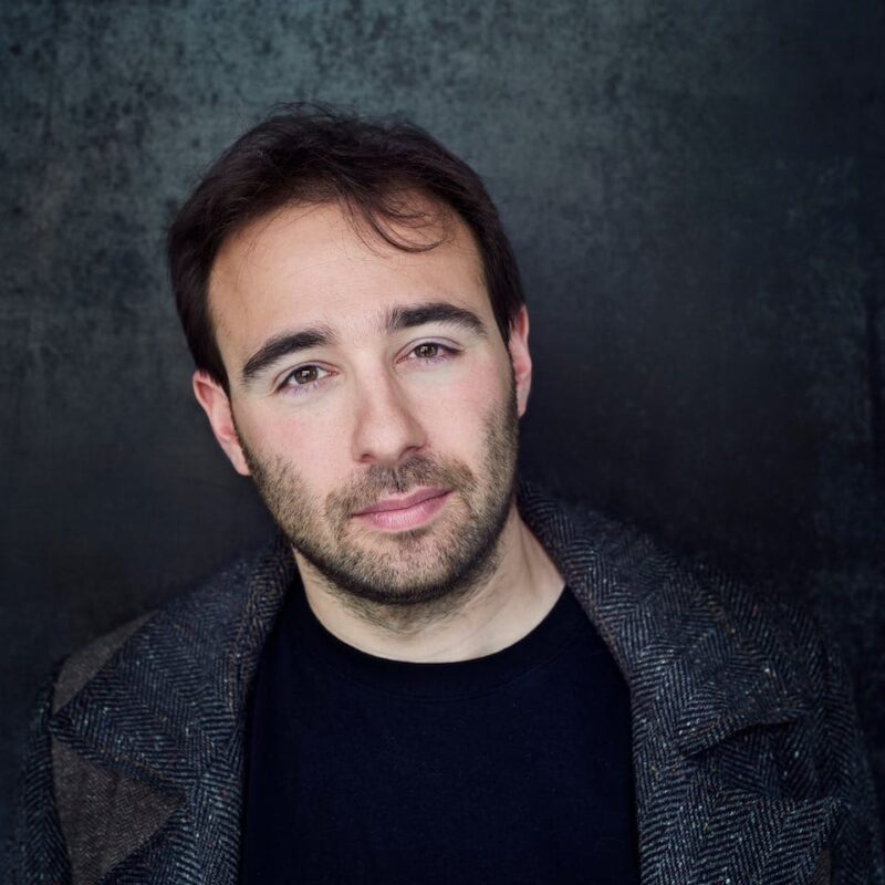 Man with short hair and a beard, wearing a dark coat, stands against a dark textured backdrop.