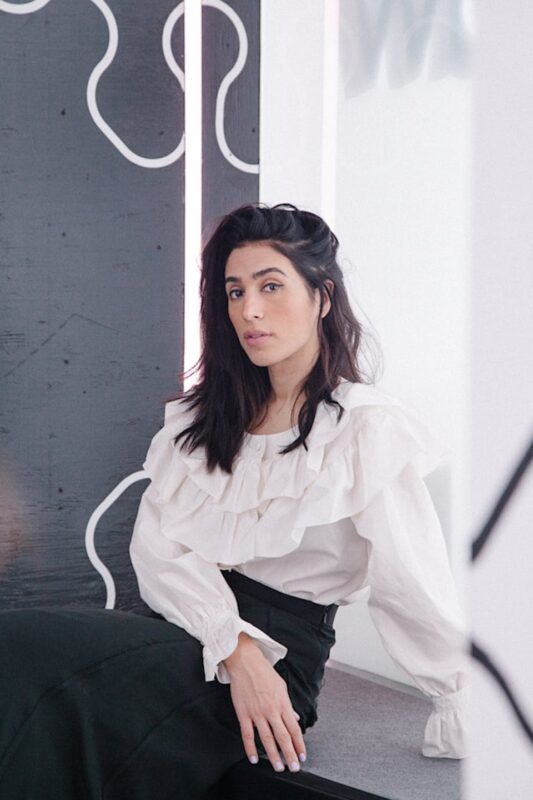 Woman in a white ruffled blouse and black skirt sits in a modern setting with abstract black and white designs in the background.