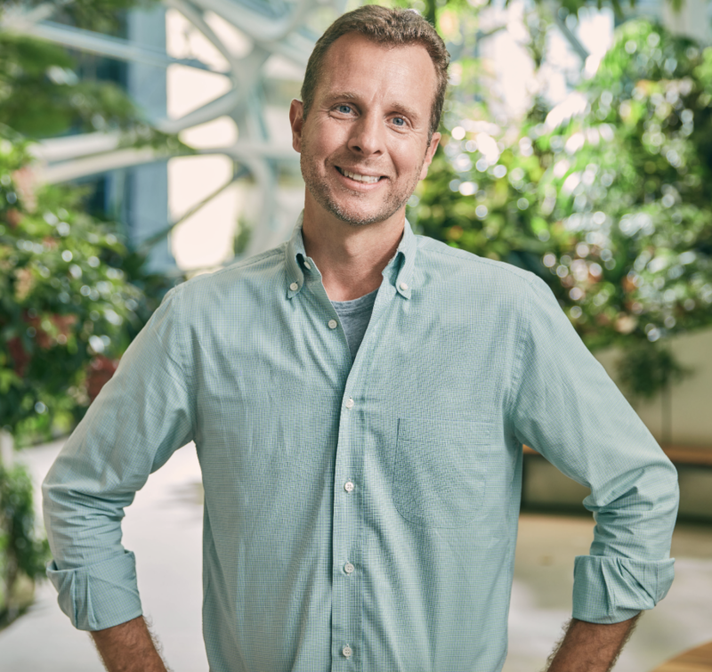 Smiling man in a green shirt stands in a bright, plant-filled environment with his hands on his hips.