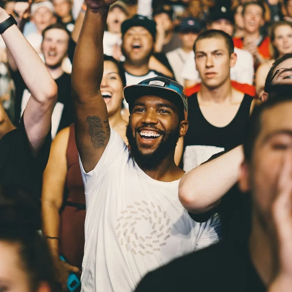 A joyful crowd at a concert, with a man in the center wearing a white Builders shirt and cap, smiling and raising his hand.