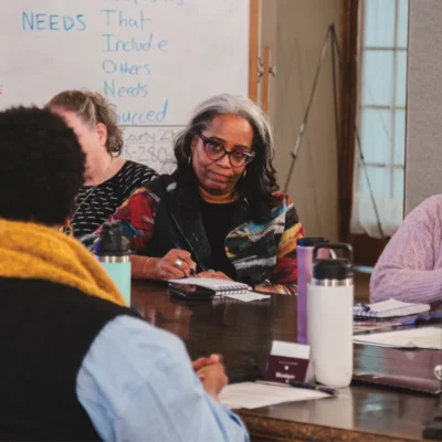 A woman with glasses and graying hair sits at a table, listening intently. Others are seated nearby, with a whiteboard in the background.