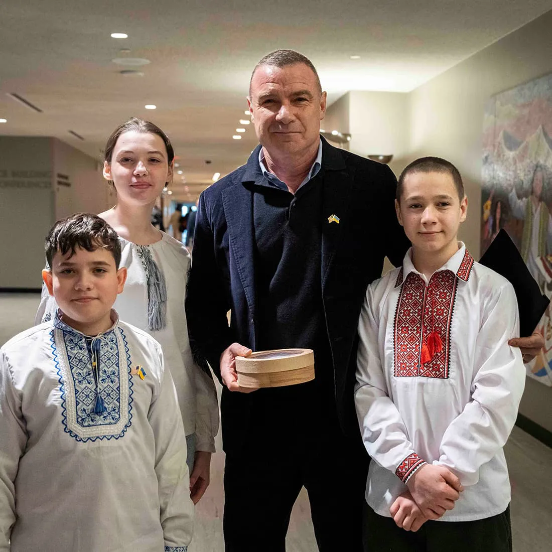 A man in a suit stands with three children wearing traditional embroidered shirts, posing for a photo indoors.